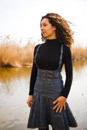 Ligia stands by a lake, dressed in a black blouse and jeans skirt.